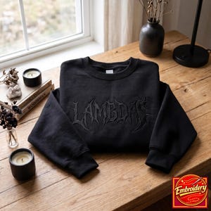 Op de afbeelding: Een zwart sweatshirt met lange mouwen en een ronde hals, voorzien van het woord LAMBDAS geborduurd in een donker, gestileerd, grillig lettertype op de borst. Het kledingstuk heeft een geribbelde kraag, boorden en zoom met ton-sur-ton stiksels.