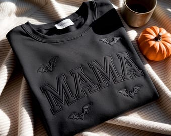 May include: Black sweatshirt featuring the word "MAMA" in raised lettering, with embroidered bat designs. A small orange pumpkin and a brown mug sit beside the sweatshirt on a cream blanket. The sweatshirt has a tag at the neckline.