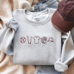 May include: A grey sweatshirt with a red embroidered design of a baseball, bats, a jersey, a glove, and a baseball cap.