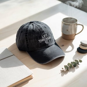 May include: A black denim baseball cap with a white embroidered message that reads "Lift Through It."