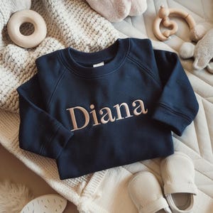 May include: A navy blue baby sweatshirt embroidered with the name "Diana" in a light pink script. The image includes baby accessories such as a wooden ring toy and soft booties.