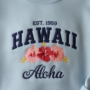 May include: Light blue sweatshirt with embroidered text "EST. 1959 HAWAII Aloha" and floral accents.