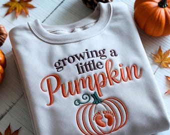 Może przedstawiać: Kremowa bluza z haftowanym napisem "growing a little Pumpkin" w kolorze brązowym i pomarańczowym. Pod tekstem znajduje się wzór dyni ze śladami stóp dziecka. Jesienna dekoracja.