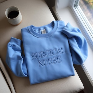 Peut inclure: Un sweat-shirt bleu clair avec le texte brodé "SURGICAL NURSE" est présenté sur une surface beige. Une tasse en céramique avec du café est à gauche. Le sweat-shirt est plié, les manches s'étendant vers l'extérieur.