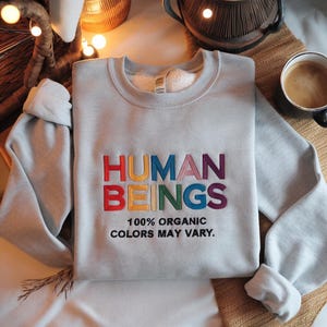May include: A light gray sweatshirt with the words "HUMAN BEINGS" embroidered in rainbow colors. The text "100% ORGANIC COLORS MAY VARY." is printed below the embroidered text.