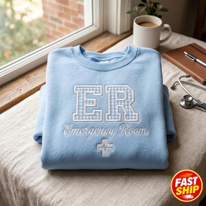 May include: Pale blue sweatshirt featuring the letters "ER" in a white and light blue gingham design, embroidered with "Emergency Room" and a medical cross. A medical theme garment.