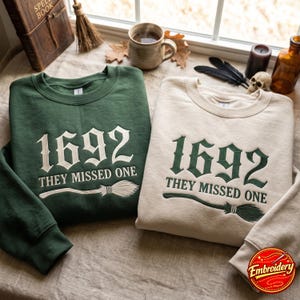 Peut inclure: Un sweat-shirt vert foncé et un sweat-shirt crème, tous deux ornés du texte brodé 1692 THEY MISSED ONE au-dessus d'un motif de balai. Le sweat-shirt vert présente une broderie blanche, tandis que le modèle crème a une broderie vert foncé.