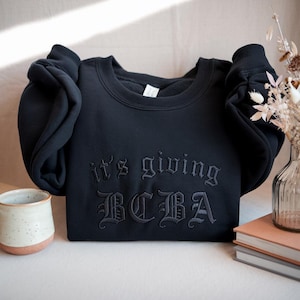May include: A black crew neck sweatshirt with the embroidered phrase "it's giving BCBA" in a gothic font. The sweatshirt is folded on a light surface, with a ceramic mug and books in the background.