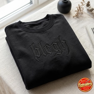May include: A black crewneck sweatshirt featuring the word blegh embroidered in a matching black thread across the chest. The garment has a ribbed collar and long sleeves, with the text rendered in a stylized, gothic-inspired font.