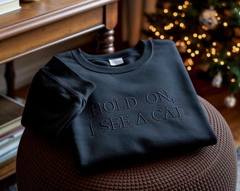 Può includere: Una felpa grigio scuro con la scritta ricamata "HOLD ON, I SEE A CAT." La felpa girocollo è piegata su un pouf marrone. Un tavolo di legno e un albero di Natale sono sullo sfondo.