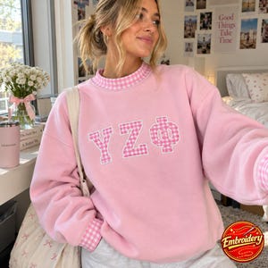 May include: A light pink crewneck sweatshirt featuring a white and pink gingham pattern on the collar, cuffs, and the Greek letters YZΦ appliqued on the chest. The sweatshirt has long sleeves and a relaxed fit, showcasing a soft fabric texture.