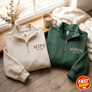 May include: Two quarter-zip sweatshirts, one beige and one green, are displayed. The beige sweatshirt has "WIFE EST. 2026" embroidered on the chest. The green sweatshirt has "HUSBAND EST. 2026" embroidered on the chest. A coffee mug and decorative vase are in the background.