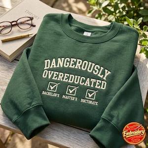 以下が含まれることがあります： クリーム色の刺繍で「DANGEROUSLY OVEREDUCATED」と書かれたフォレストグリーンのクルーネックトレーナー。テキストの下にはチェックマークが入った3つのチェックボックスがあり、それぞれ「BACHELOR'S」「MASTER'S」「DOCTORATE」とクリーム色の文字で記されています。