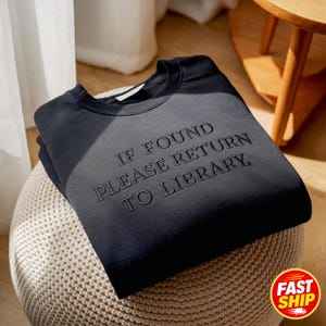 May include: A black crewneck sweatshirt featuring the embroidered text IF FOUND PLEASE RETURN TO LIBRARY in a matching black thread. The garment has long sleeves, a ribbed neckline, and a clean, solid-colored design with a subtle texture.