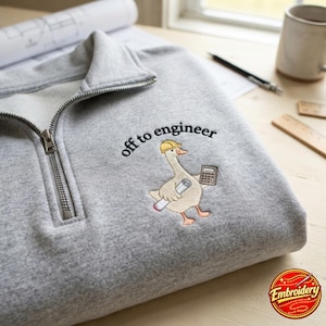 Puede incluir: Una sudadera gris claro con cremallera corta que presenta un diseño bordado de un ganso con casco amarillo, sosteniendo un plano enrollado y una calculadora, con el texto bordado en negro "off to engineer" situado sobre el ganso.