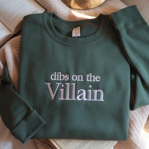 May include: A dark green sweatshirt with the embroidered phrase "dibs on the Villain" in white. The sweatshirt is folded on a neutral-toned blanket, with a book visible in the background. The sweatshirt has a crew neck and long sleeves.