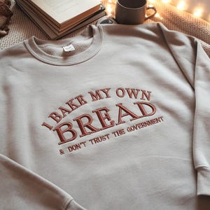 Peut inclure: Un sweat-shirt gris clair avec le texte "I bake my own bread & don't trust the government" brodé en rouge.