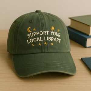 May include: A green baseball cap with the embroidered text "SUPPORT YOUR LOCAL LIBRARY" in white. The design includes a yellow crescent moon, sun, and stars. The cap is sitting on a wooden surface.