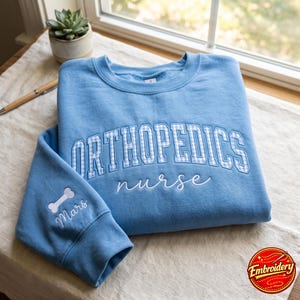 Könnte beinhalten: Ein hellblaues Sweatshirt mit Rundhalsausschnitt und dem gestickten Text ORTHOPEDICS in blau-weißem Vichy-Muster, darunter das Wort nurse in weißer Schreibschrift. Der Ärmel zeigt ein gesticktes weißes Knochensymbol und den Namen Mars.