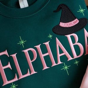 Pu&ograve; includere: Felpa verde scuro con la scritta ricamata "ELPHABA" in rosa, un cappello da strega nero con una fascia rosa e stelle verdi. Un capo casual e comodo.