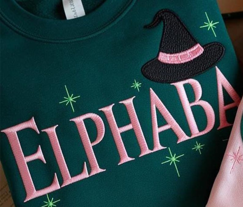 Pu&ograve; includere: Felpa verde scuro con la scritta ricamata "ELPHABA" in rosa, un cappello da strega nero con una fascia rosa e stelle verdi. Un capo casual e comodo.