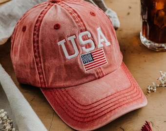 May include: A red baseball cap with a white embroidered "USA" and a small American flag on the front. The cap has a curved brim and a distressed look.