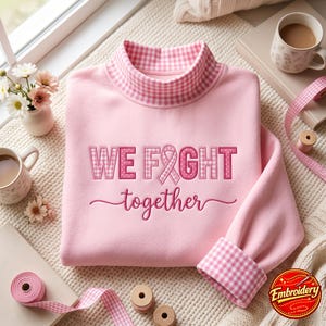 Op de afbeelding: Een lichtroze sweatshirt met een opstaande kraag in een roze en wit vichy-ruitpatroon. Op de borst staat de geborduurde tekst WE FIGHT together, waarbij de letter I is vervangen door een roze bewustzijnslint in een vichy-patroon.