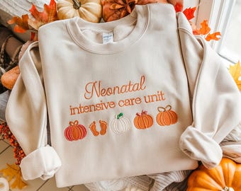 Pode incluir: Camisola creme com a frase bordada "Neonatal intensive care unit" em laranja. Abaixo, bordados de abóboras, pegadas de bebé e uma abóbora branca. A camisola é rodeada por abóboras e folhas de outono.