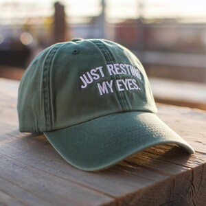 May include: A green baseball cap with the text "JUST RESTING MY EYES" embroidered in white. The cap has a curved brim and is made of a soft, worn material. The hat is sitting on a wooden surface.