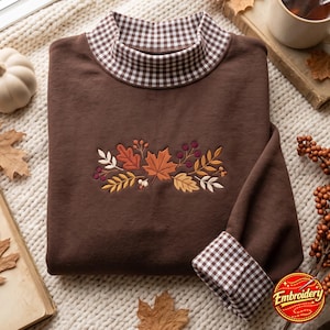Puede incluir: Una sudadera marrón de manga larga con cuello alto y puños con un patrón de cuadros vichy en marrón y blanco. El centro del pecho presenta un diseño bordado de hojas de otoño, bayas y ramitas en tonos naranja, marrón y crema.