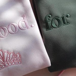 K&ouml;nnte beinhalten: Zwei Sweatshirts, eines in Hellrosa und eines in Dunkelgr&uuml;n, mit gestickten Texten und Designs. Das rosa Sweatshirt hat den Schriftzug "for good." mit einer Krone. Das gr&uuml;ne Sweatshirt hat den Schriftzug "for good." mit einem Hexenhut.