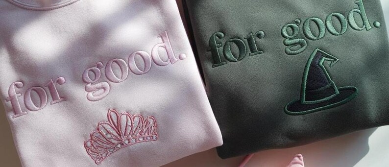 K&ouml;nnte beinhalten: Zwei Sweatshirts, eines in Hellrosa und eines in Dunkelgr&uuml;n, mit gestickten Texten und Designs. Das rosa Sweatshirt hat den Schriftzug "for good." mit einer Krone. Das gr&uuml;ne Sweatshirt hat den Schriftzug "for good." mit einem Hexenhut.
