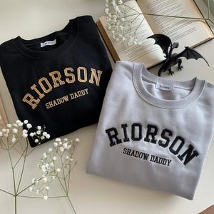 May include: Two sweatshirts with the text "RIORSON SHADOW DADDY" in black and gold embroidery. One sweatshirt is black and the other is gray.