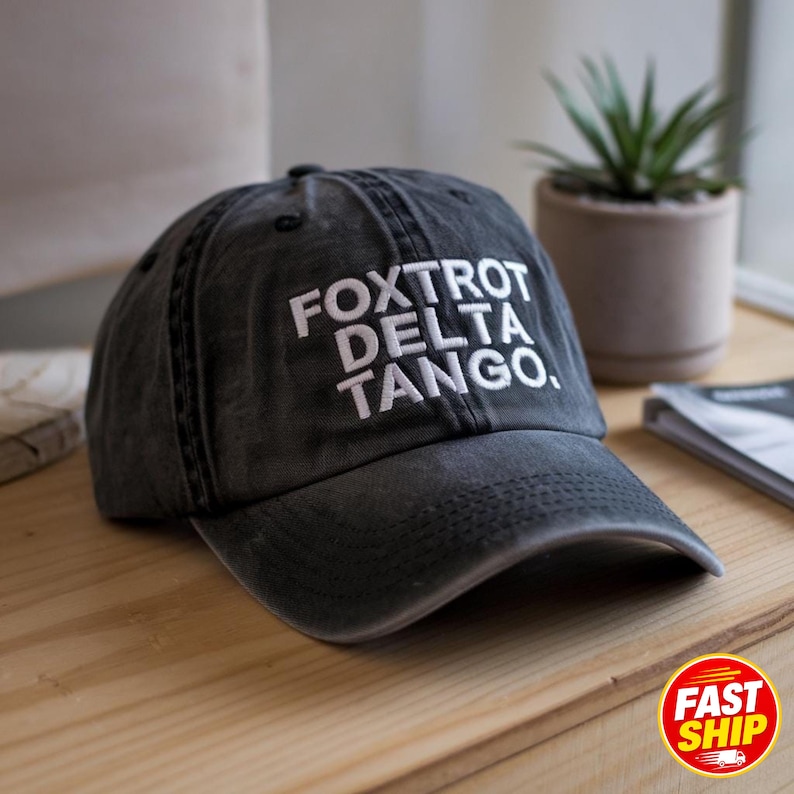 May include: A charcoal gray baseball cap with a distressed, washed-out finish. The front of the cap features white embroidered text that reads FOXTROT DELTA TANGO. The hat has a structured crown, a curved brim, and visible stitching details.