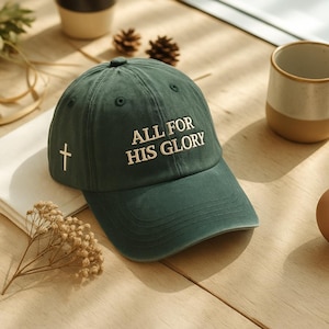 May include: A dark green baseball cap with the text "ALL FOR HIS GLORY" embroidered in white. A white cross is embroidered on the side. The cap has a curved brim and a button on top.