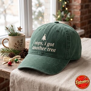 May include: A forest green baseball cap featuring an embroidered white Christmas tree icon and the text "oops, i got another tree" in a white serif font. The cap has a structured crown, a curved brim, and stitched eyelets on the panels.