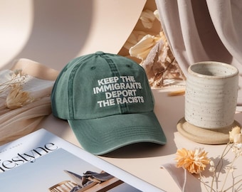 May include: A green baseball cap with the text "KEEP THE IMMIGRANTS, DEPORT THE RACISTS" in white. The cap is on a table next to a magazine and a ceramic mug.