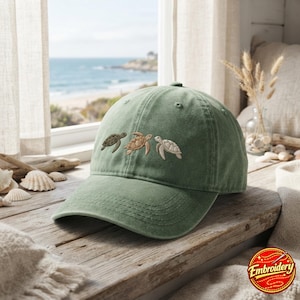 May include: A sage green baseball cap featuring an embroidered design of three sea turtles in varying shades of brown and cream. The cap has a structured crown, a curved brim with visible stitching, and eyelets for ventilation on the top panels.