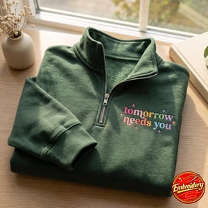 Puede incluir: Una sudadera verde oscuro con cremallera de un cuarto, cierre metálico plateado y puños acanalados. En el pecho presenta el texto bordado "tomorrow needs you" en un degradado multicolor, decorado con pequeñas estrellas.