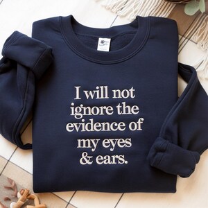 May include: Navy blue sweatshirt with the quote "I will not ignore the evidence of my eyes & ears" in white lettering. The sweatshirt has a crew neck and long sleeves, folded at the wrist.