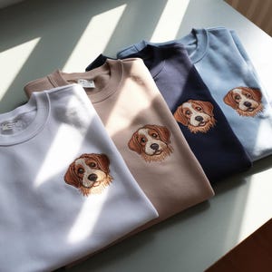 May include: Four sweatshirts in different colors with embroidered dog faces. The sweatshirts are light blue, navy blue, beige, and white.
