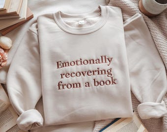 May include: A cream-coloured sweatshirt with the embroidered text "Emotionally recovering from a book" in a brown font. The sweatshirt is on a beige blanket, with open books and a mug nearby.