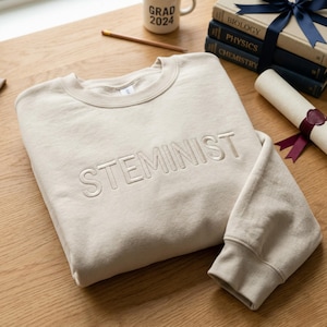 Könnte beinhalten: Beiges Sweatshirt mit dem Wort "STEMINIST" auf der Vorderseite gestickt. Das Sweatshirt liegt gefaltet auf einer Holzoberfläche, mit einer "Grad 2024"-Tasse, Büchern, einem Bleistift und einem Diplom in der Nähe. Das Sweatshirt hat einen Rundhalsausschnitt und gerippte Bündchen.