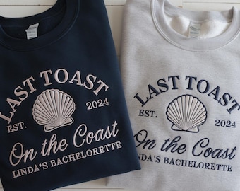 Op de afbeelding: Twee sweatshirts, een donkerblauwe en een lichtgrijze, met de tekst "Last Toast" in een witte boog boven een witte schelp. De tekst "EST. 2024" staat onder de schelp. De tekst "On the Coast" staat onder de schelp en de tekst "Linda's Bachelorette" staat daaronder.