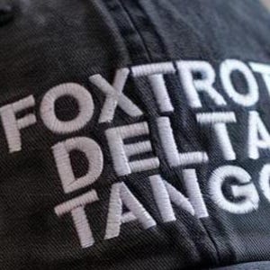 May include: A charcoal gray baseball cap featuring the white embroidered text FOXTROT DELTA TANGO. The cap has a visible fabric texture and a structured crown with a button at the top, showcasing clear stitching details on the fabric panels.