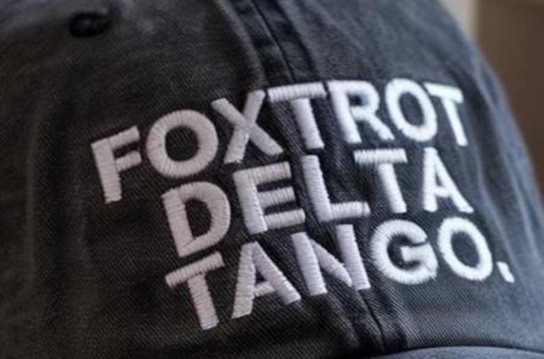 May include: A charcoal gray baseball cap featuring the white embroidered text FOXTROT DELTA TANGO. The cap has a visible fabric texture and a structured crown with a button at the top, showcasing clear stitching details on the fabric panels.