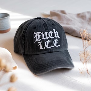 May include: A dark grey baseball cap featuring the white embroidered text "Fuck I.C.E." in a gothic font. The cap has a pre-worn look and a curved peak. A casual hat.