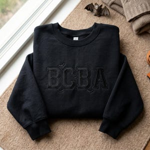 May include: A black crewneck sweatshirt featuring the letters BCBA embroidered in a matching black thread on the chest. The design includes subtle embroidered details of small bats and spiderwebs surrounding the bold, block-style lettering.