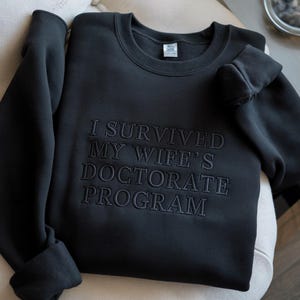 May include: Black crewneck sweatshirt with the embroidered text "I SURVIVED MY WIFE'S DOCTORATE PROGRAM". The sweatshirt is made of a soft material and has long sleeves. The text is in a darker shade of black.