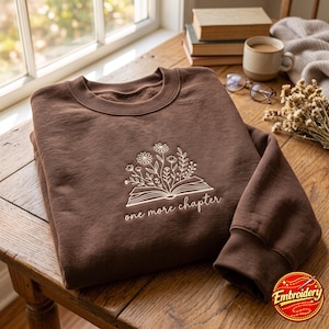 Pode incluir: Uma camisola de gola redonda castanho-escura com um design bordado de um livro aberto com flores a crescer das páginas. Por baixo do livro, o texto "one more chapter" está bordado numa fonte cursiva de cor creme claro.